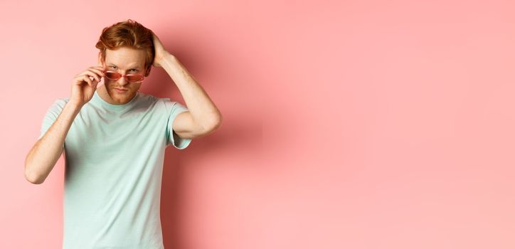 Handsome young redhead man in sunglasses, brushing hair with hand and looking smug and confident at camera, standing over pink background.