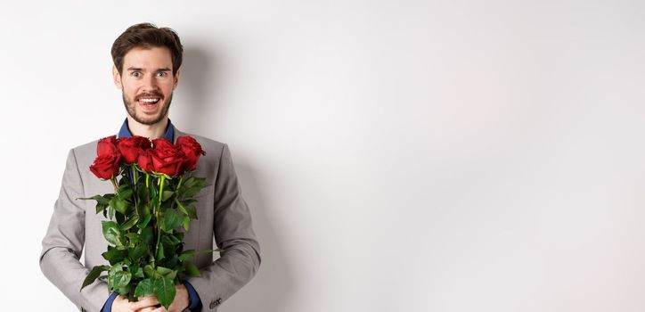 Excited handsome man in suit holding bouquet of roses for romantic date with lover, standing happy on Valentines day, white background.