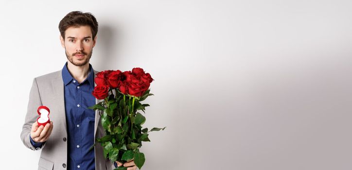 Handsome man in suit going to make a proposal, standing with red roses flowers and engagement rin in box, making romantic surprise, white background.