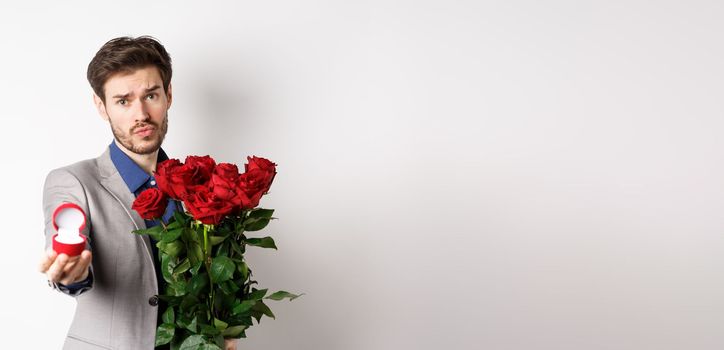 Handsome boyfriend in suit asking to marry him, standing with red bouquet of roses and engagement ring, looking pleading at camera, standing over white background.