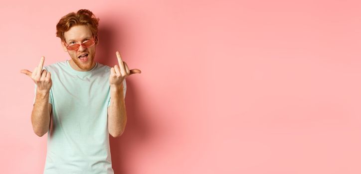 Arrogant and careless redhead man in sunglasses dont give a fuck, showing middle fingers at camera and frowning, standing over pink background.