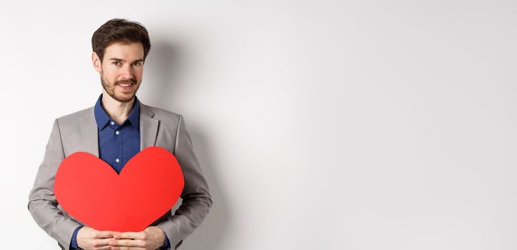 Handsome bearded man wishing happy valentines day, holding red heart cutout and smiling, going on romantic date in fancy suit, standing over white background.