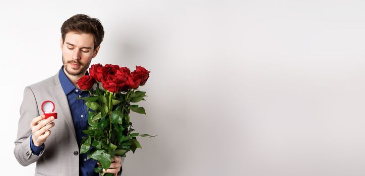 Romantic man in suit looking pensive at engagement ring, going to make a marriage proposal on Valentines day, holding bouquet of roses, standing over white background.