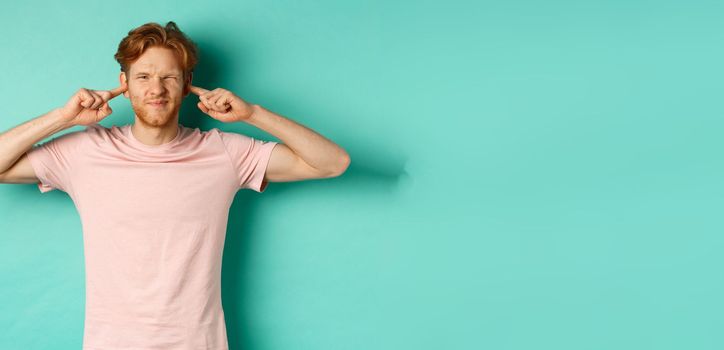 Annoyed young man with red hair and beard shut his ears and grimacing, disturbed by loud bothering sound, noisy neighbours, standing over turquoise background.