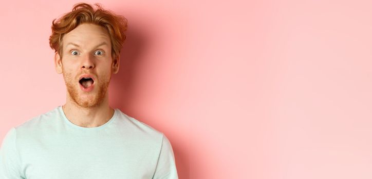 Face of surprised redhead man reacting to cool promo offer, raising eyebrows and gasping, staring in awe at camera, standing over pink background.