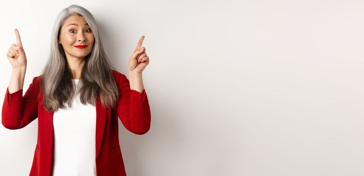 Elegant asian senior lady in red blazer and makeup, pointing fingers up and smiling, showing advertisement, white background.