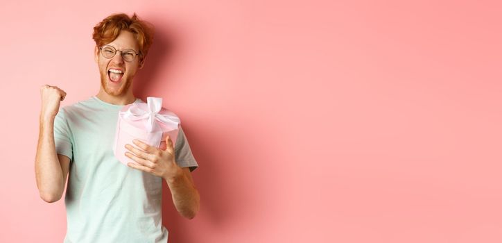 Valentines day and romance concept. Cheerful redhead guy making fist pump and scream yes with joy, holding gift box, smiling, standing over pink background.