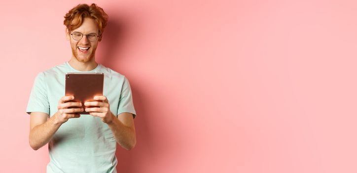 Happy guy with red hair and beard using digital tablet, reading screen and smiling, standing over pink background.