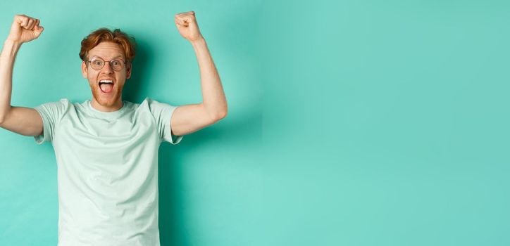 Young redhead man feeling like champion, raising hands up in fist pump gesture and shouting yes with joy, winning prize, triumphing of success, standing over mint background.