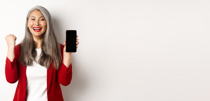 Success. Asian senior woman showing smartphone blank screen and fist pump, winning prize online, standing over white background.