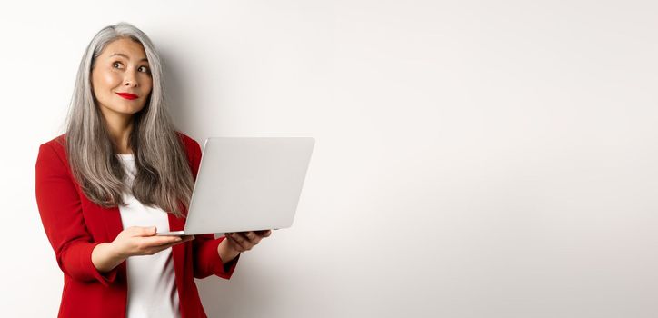Business. Successful asian businesswoman in red blazer imaging something, working on laptop and looking upper left corner with dreamy smile, standing over white background.