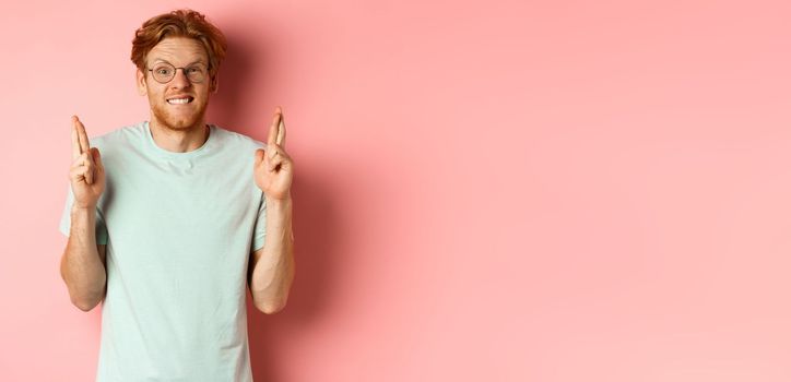 Worried redhead man waiting for results, expecting something with fingers crossed, biting finger and looking at something risky, standing over pink background.