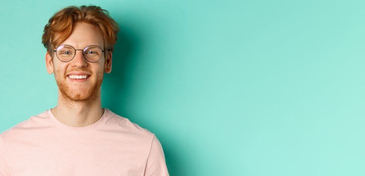 Close up of handsome redhead man in glasses looking at camera, smiling with white teeth, standing against mint background.