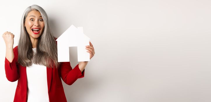Happy asian grandmother showing paper house cutout and fist pump gesture, scream yes with joy, buying property, standing over white background.