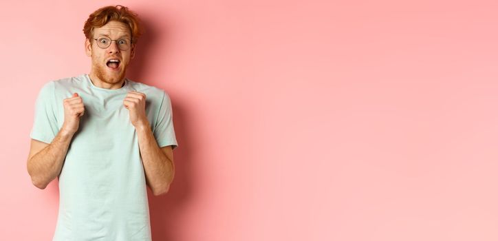 Portrait of scared redhead guy in glasses staring startled at camera, press hands to body and scream of fear, standing over pink background.