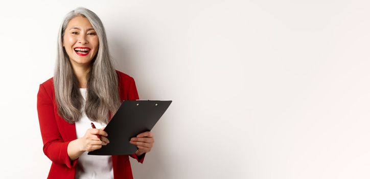 Happy asian office employee, woman in red blazer, working and laughing, holding clipboard with documents, standing over white background.