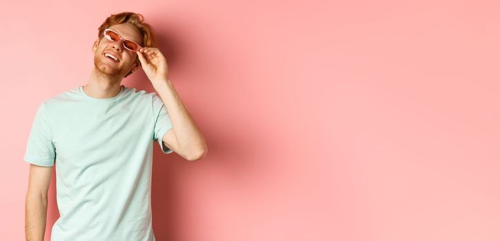 Tourism and vacation concept. Happy young man with red hair relaxing, wearing sunglasses and smiling carefree, standing over pink background.