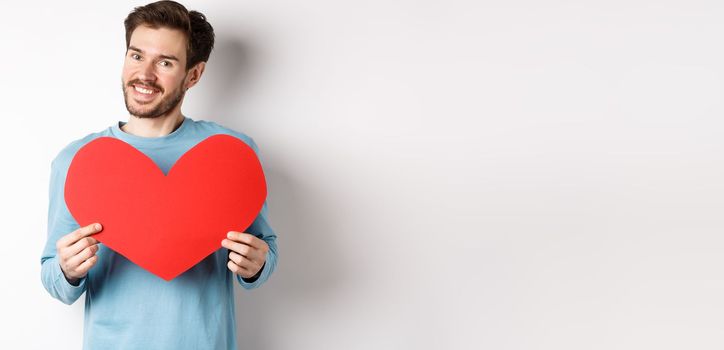 Handsome european man in sweater saying I love you, boyfriend standing with valentines day red heart, posing over white background.