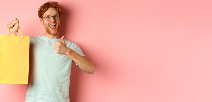 Cheerful redhead man in t-shirt and glasses pointing finger at shopping bag, showing shop with discounts, standing over pink background.