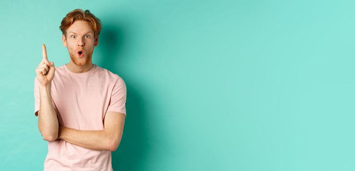 Pensive redhead man in t-shirt raising index finger, gasping as pitching at idea, saying suggestion, standing over turquoise background. Copy space