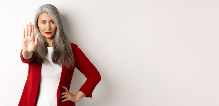 Serious asian woman in red blazer showing stop sign, telling no, frowning and rejecting something bad, standing against white background.