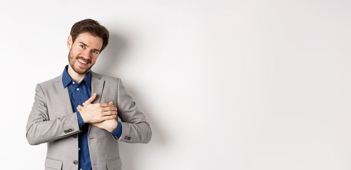 Happy man in suit holding hands on heart, feeling touched or grateful, having tender memories in soul, standing against white background.