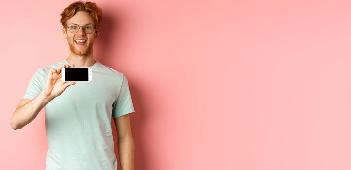 Happy young man with red hair showing smartphone screen, holding phone horizontally and smiling amazed, standing over pink background.