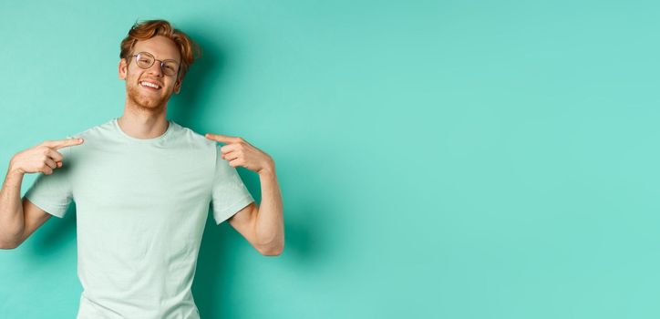 Confident redhead man in glasses and t-shirt, smiling with smug face and pointing at himself, bragging while standing over turquoise background.