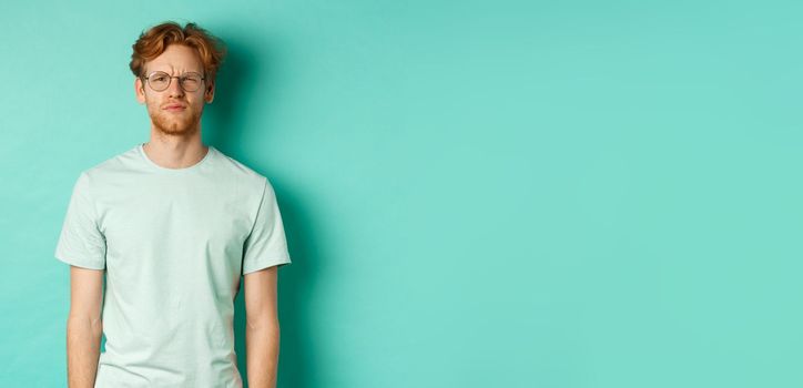 Confused young redhead man in glasses staring at camera puzzled, squinting perplexed, cant understand something, standing over mint background.