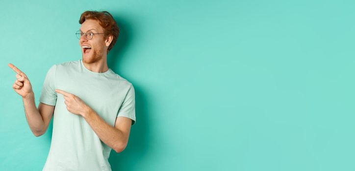 Excited young man with red hair, wearing glasses and t-shirt, pointing and looking left at awesome promotion, smiling happy at banner, turquoise background.