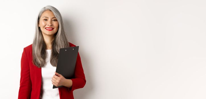 Successful asian senior business woman holding clipboard, wearing red blazer and lipstick at work, smiling at camera, white background.