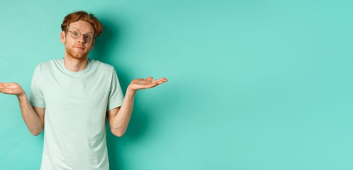 Portrait of confused redhead man in t-shirt and glasses know nothing, shrugging shoulders and looking clueless at camera, standing against turquoise background.