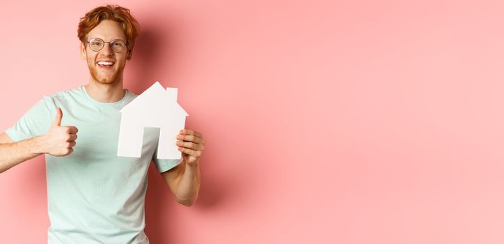 Real estate. Cheerful man in glasses and t-shirt recommending broker agency, showing paper house cutout and thumbs-up, standing over pink background.