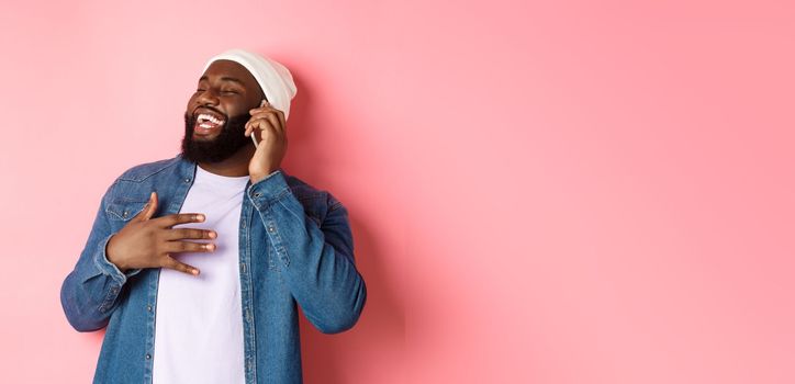 Happy african-american man talking on mobile phone, laughing and smiling, standing in beanie and denim shirt over pink background.