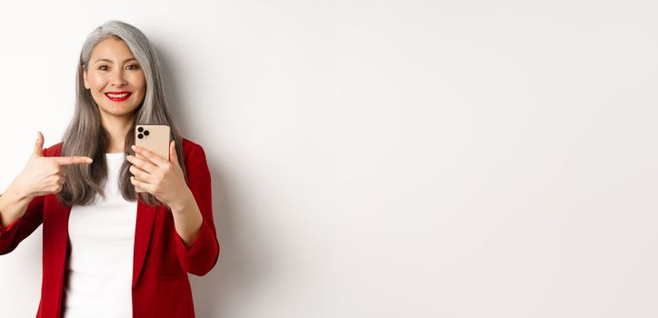 Asian elderly woman in elegant blazer showing smartphone, pointing finger at mobile phone and smiling, standing over white background.