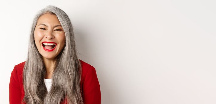 Close-up of happy asian businesswoman with long grey hair, wearing red blazer, laughing and smiling joyfully at camera, white background.