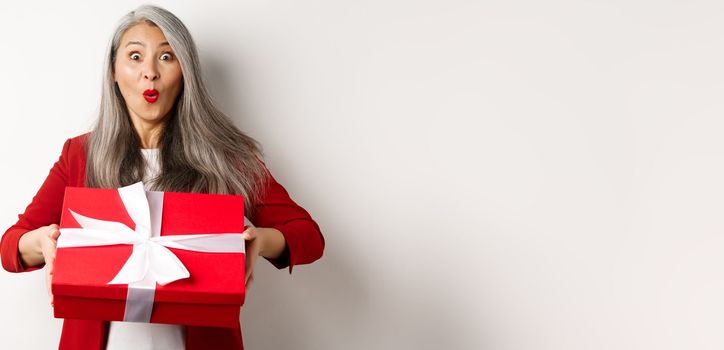 Surprised asian senior woman receiving present on mother day, holding red box with gift and looking amazed at camera, white background.