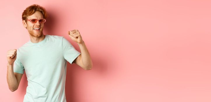 Tourism and vacation concept. Cheerful redhead man having fun at party, dancing and enjoying holiday, standing in sunglasses and t-shirt against pink background.