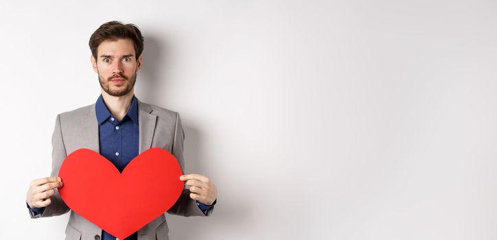 Excited caucasian man in suit looking at camera, holding big red heart cutout on valentines day, standing over white background. Copy space