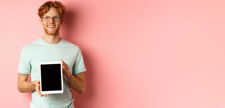 Funny redhead guy in glasses demonstrate digital tablet screen, smiling at camera, standing over pink background.