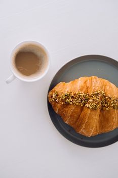 Flat lay of cup of cappuccino with pistachio croissant on white table