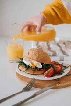Breakfast sandwich with egg and arugula and cherry tomatoes on table, woman pours orange juice into a glass