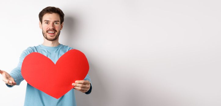 Handsome man in love making confession to you, pointing hand at camera, holding big red heart cutout on valentines day, singing romantic serenade, standing over white background.