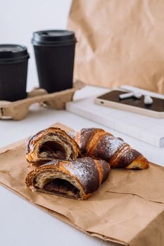 Chocolate croissants with cups of coffee and notepad with smartphone and earphones on white table, food delivery concept