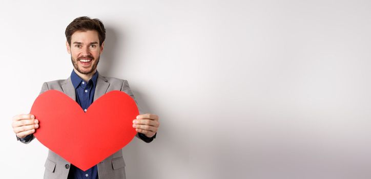 Handsome young man wishing happy Valentines day, giving big red heart sign and smiling, make surprise to lover, standing in suit over white background.