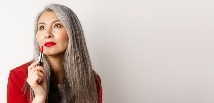 Beauty and makeup concept. Sensual mature asian female with grey hair, looking aside and showing red lipstick, standing over white background.