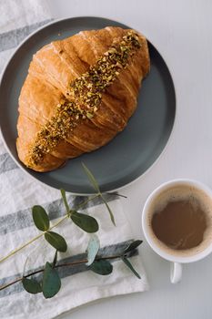 Flat lay of cup of cappuccino with kitchen towel, croissant and eucalyptus branch on table