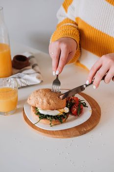 Unrecognizable woman eating breakfast sandwich with egg and arugula and cherry tomatoes on table with orange juice