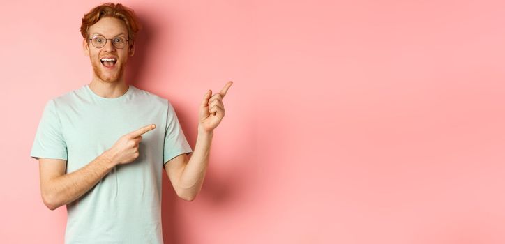 Amazed redhead man in glasses and t-shirt showing special promotion, pointing fingers at upper right corner and staring excited at camera, standing over pink background.