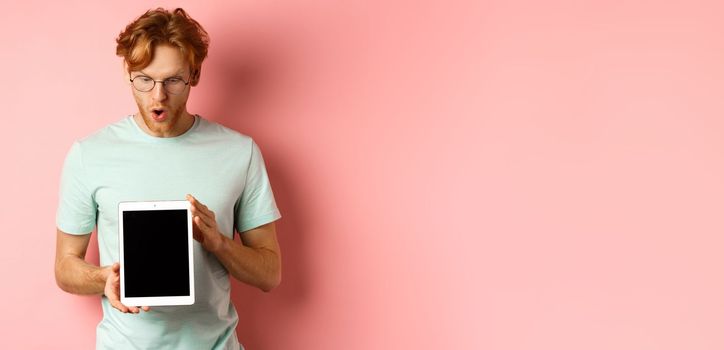Impressed redhead man in glasses, showing blank digital tablet screen and looking in awe at display, saying wow, standing in t-shirt against pink background.